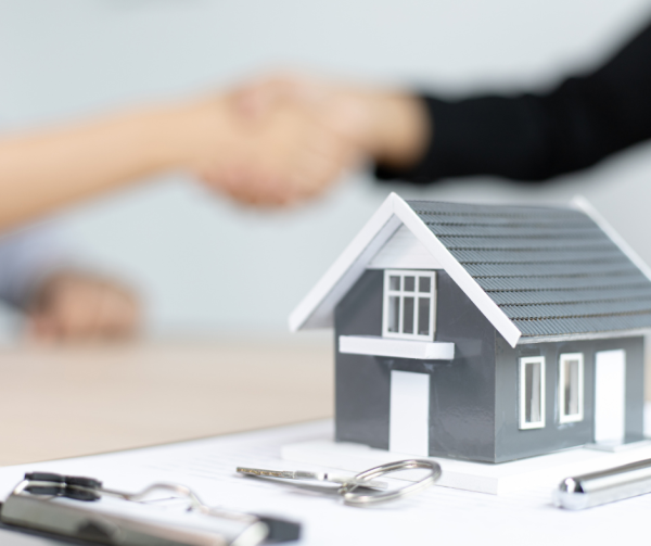 Image shows a small model house sitting on a clipboard on a desk with a key in front of it. Behind, blurred, is two people shaking hands.