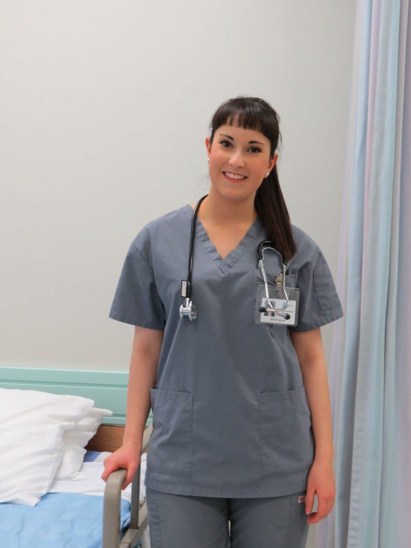 Image of young woman with stethoscope, in scrubs, standing by hospital bed.