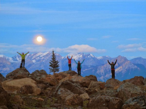 Image shows four individuals with their arms raised on top of a mountain with the moon behind them.