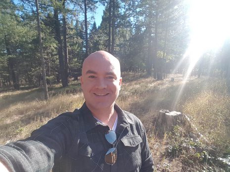 Image shows man smiling out in the woods with bright sunlight behind him.