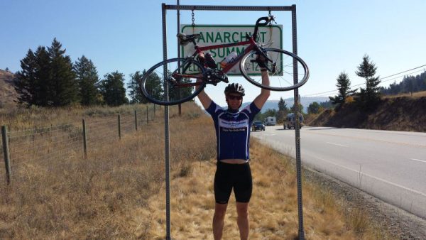 Image of man holding a bike over his head next to a highway and in front of a sign which says Anarchist Summit.