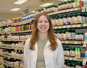 Image shows woman in white medical coat standing in front of a aisle with many medications.