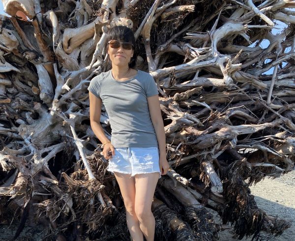 Image shows woman in sunglasses in front of the roots of a large overturned tree, smiling at the camera.