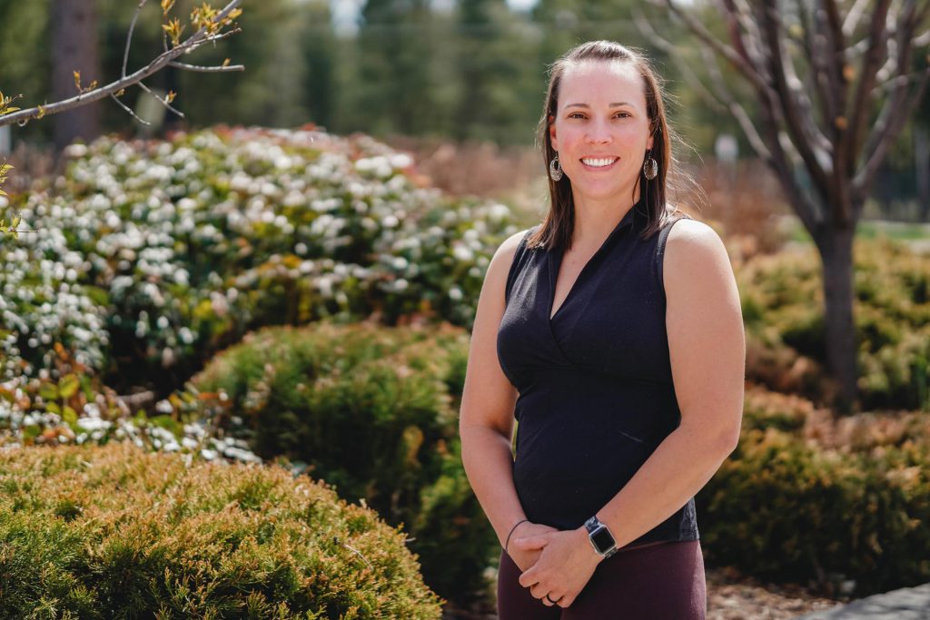 Image shows woman smiling outside with shrubs, bushes and trees around her.