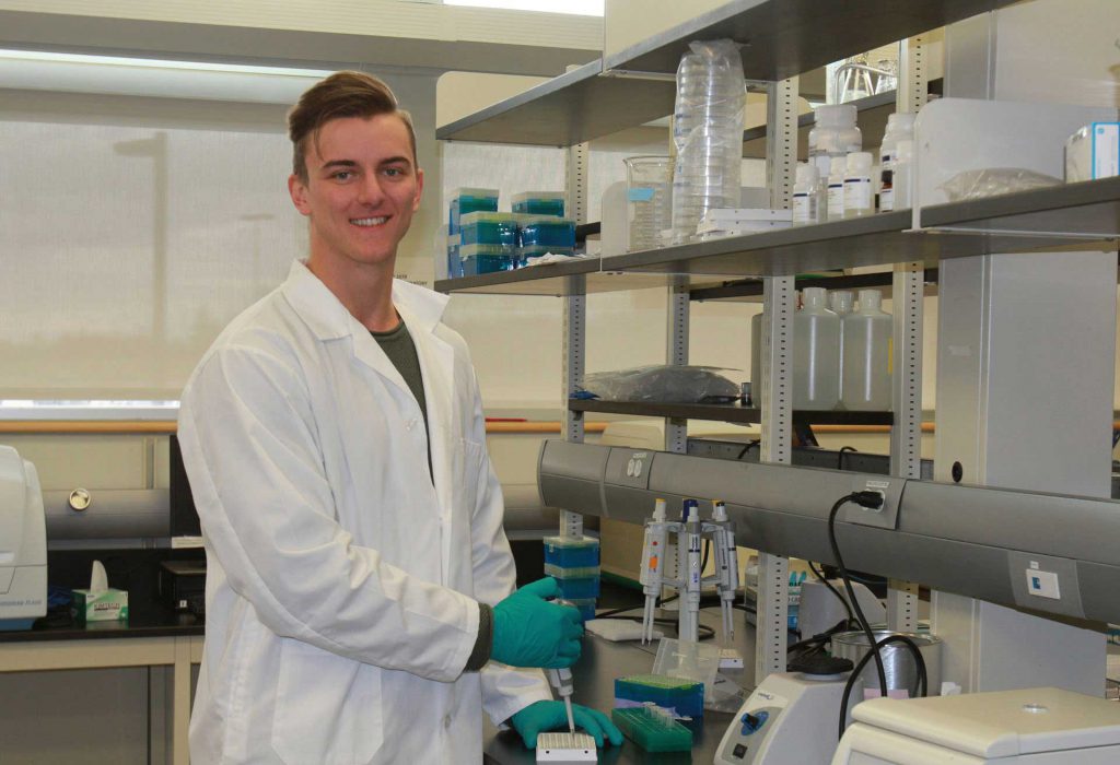 Image shows man in white lab coat and gloves in a lab setting.