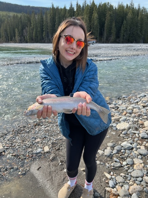 Image shows young woman in sunglasses, next to a river, holding a fish