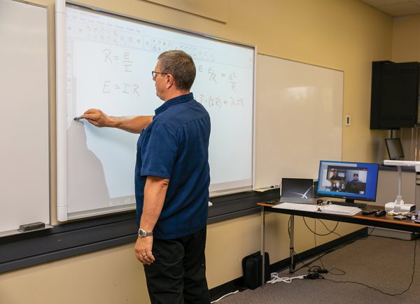 Image shows male instructor writing on a smart board with his students looking on via computer.