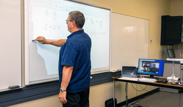 Image shows man standing writing on a Smartboard while students watch via a nearby computer.