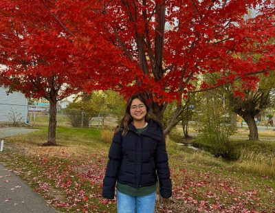 Image shows young woman standing outside in front of a maple tree with vivid red leaves