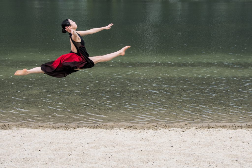 Image shows young woman leaping in a full splits in the air on a beach.