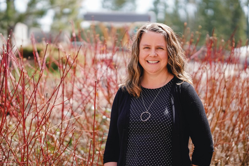 Image shows woman smiling at camera with colourful foliage behind her.