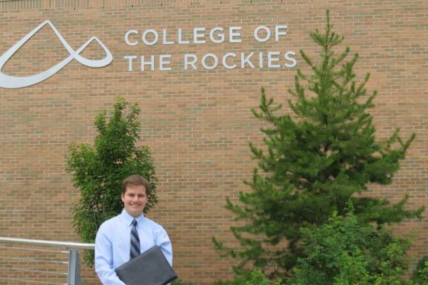 Image of young man in shirt and tie, carrying a porfolio, standing in front of College of the Rockies