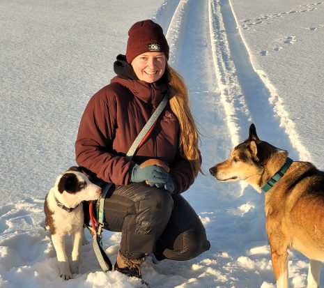 Image shows woman crouching down in the snow with her two dogs.