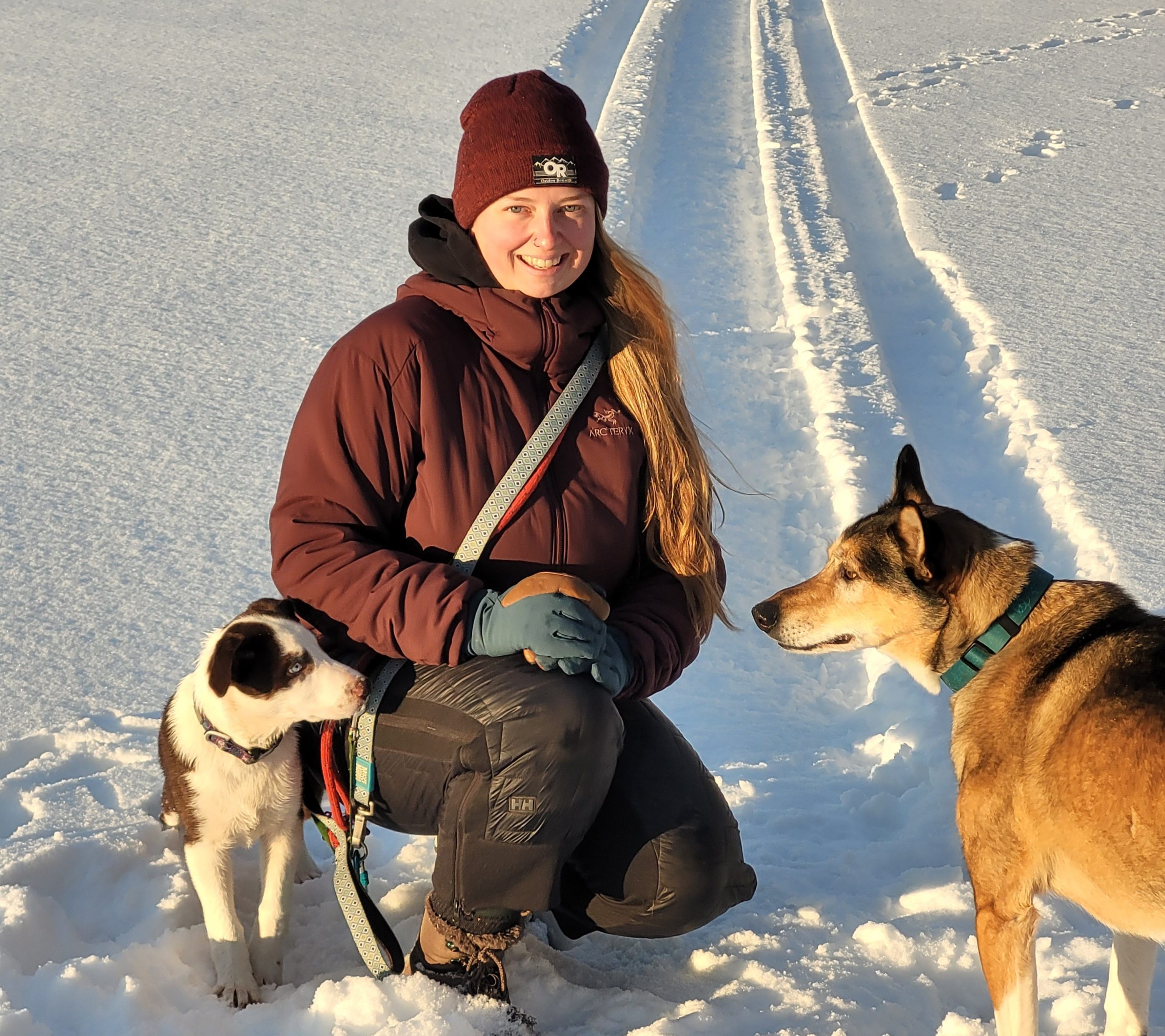 Image shows woman crouching down in the snow with her two dogs.
