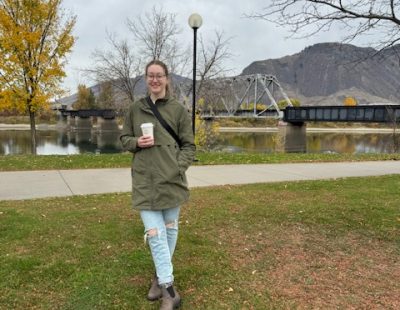 Image shows young woman with glasses standing outdoors wearing jeans, a green jacket, a pair of Blundstones, with a bag strapped around her chest and her holding a coffee cup. She is outdoors in fall with a train track and mountains behind her.