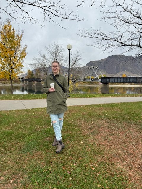 Image shows young woman with glasses standing outdoors wearing jeans, a green jacket, a pair of Blundstones, with a bag strapped around her chest and her holding a coffee cup. She is outdoors in fall with a train track and mountains behind her.