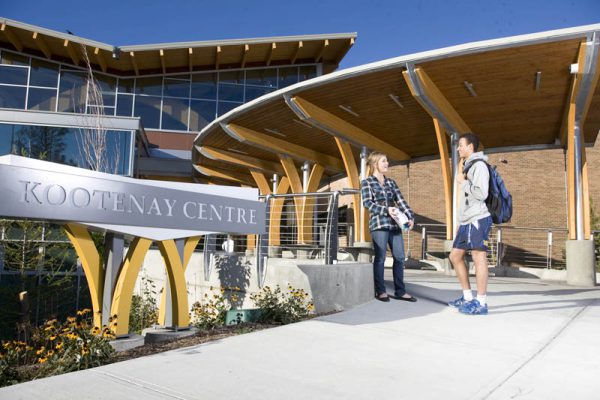 Image shows front entrance of College of the Rockies with two students standing by Kootenay Centre sign.