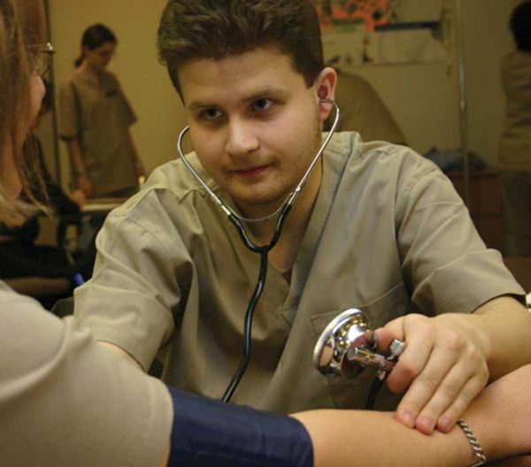 Image of male in scrubs, with stethoscope in ears, taking blood pressure of an individual.
