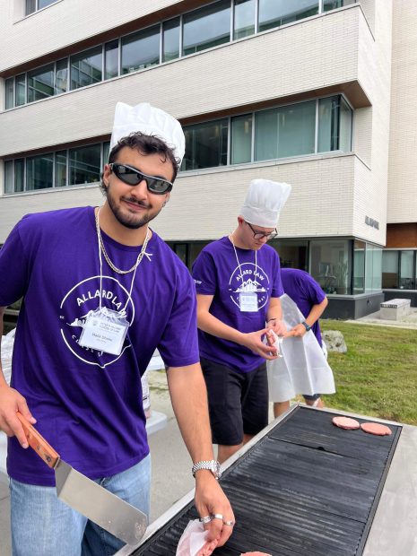 Image shows man in purple shirt and chef's hat BBQ with others.