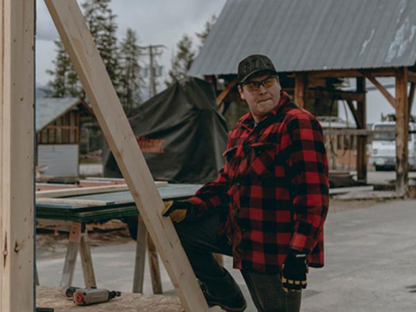 Image of man with red and black checked jacket, and work gloves standing next to a carpentry project.