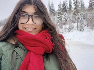 Image shows woman with long brown hair and glasses standing otudoors in the winter wearing a heavy jacket and a red scarf