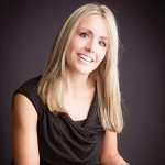 Portrait of a blonde woman wearing a flattering black top in front of a black background.