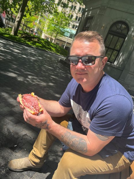 Image shows man in sunglasses sitting outdoors, holding a very large sandwich.