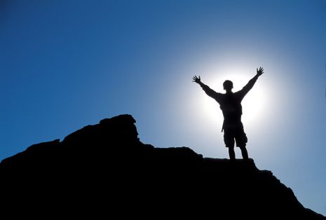 Image shows silhouette of a person standing, with arms raised, on the top of a mountain.