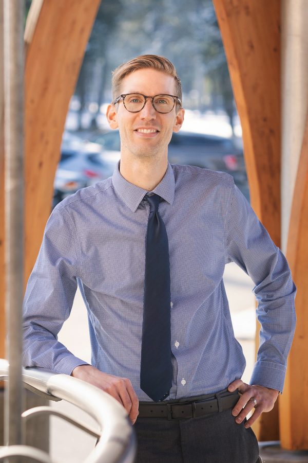 Image shows man in dress shirt and tie, wearing glasses, leaning against a railing and smiling at the camera.