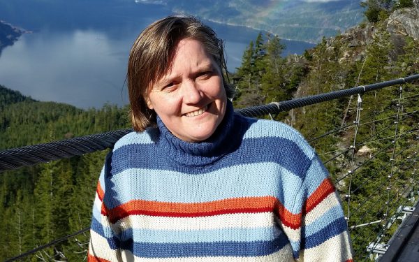 Image shows smiling woman in a striped sweater with trees and a lake behind her.