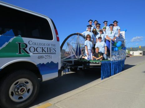 Image of a group of 13 people on a parade float, some wearing graduation hats, being pulled by a College of the Rockies van.
