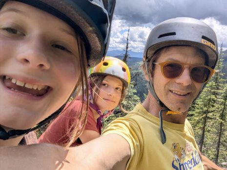 Image shows man and two kids wearing bike helmets and smiling at the camera.