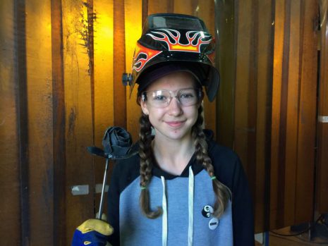 Image shows young girl in safety glasses and welding mask holding a fabricated rose.