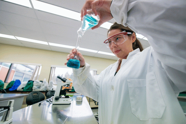 Image of young woman in white lab coat and safety glasses pouring blue liquid from one beaker to another.