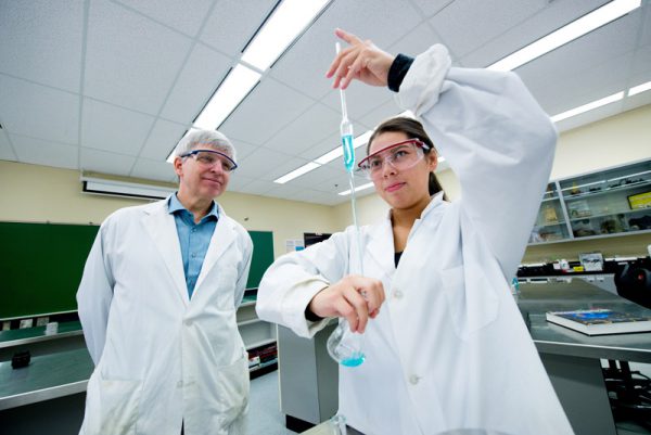 Image of young woman in white lab coat at safety goggles transferring liquids between beakers while a grey-haired man looks on.