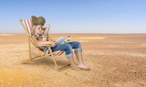Image shows young woman reading while sitting on a beach chair surrounded by sand.