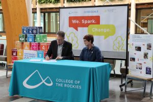 Image shows two people at a table with a College of the Rockies tablecloth, with the words We. Spark. Change. behind them and blocks representing the 17 Sustainable Development Goals .