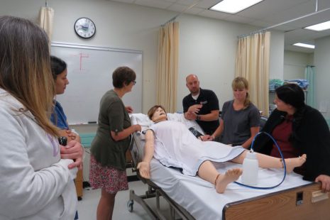 Image shows students and instructors gathered around a simulator mannequin lying on a hospital bed.