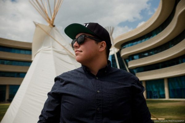 Image shows man in a ball cap and sunglasses with a teepee and a building behind him.
