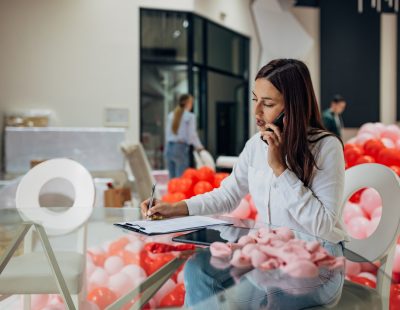 A female event planner sits in a restaurant and talks to new clients on the phone, writes down the details of the reservation and the decoration that the client wants