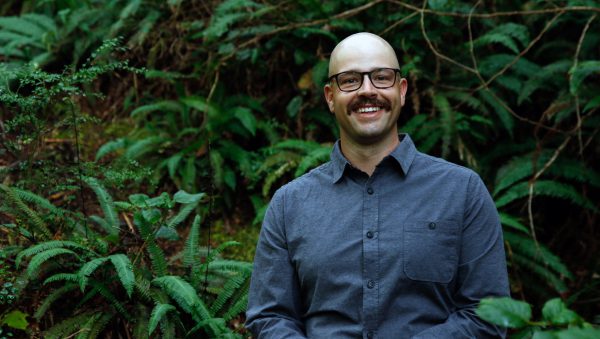 Image of man in glasses smiling at camera with lush foliage behind him.