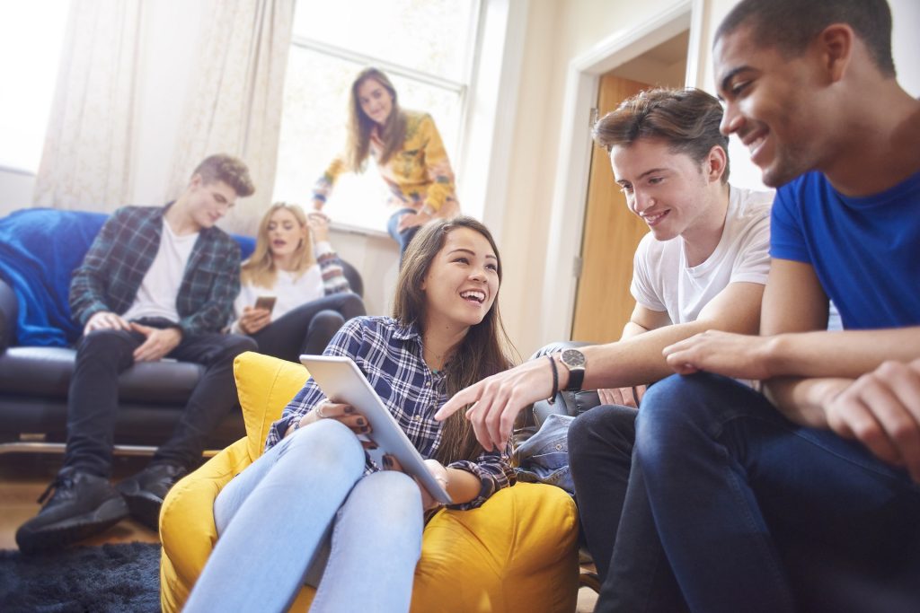 six young students sit around in their halls of residence , chatting and socialising with mobile phones and digital tablets. Three women and three men sit on sofas , beanbags and window ledges chatting and laughing .