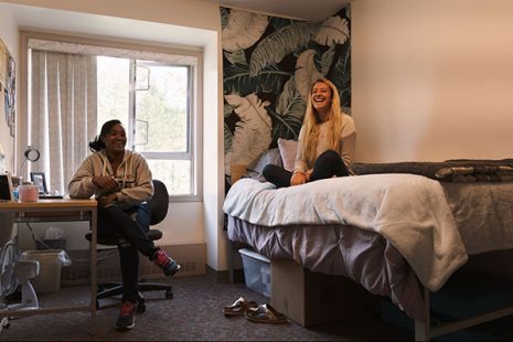 Image shows two students laughing inside a dorm room, one sitting on a chair, the other on a bed.
