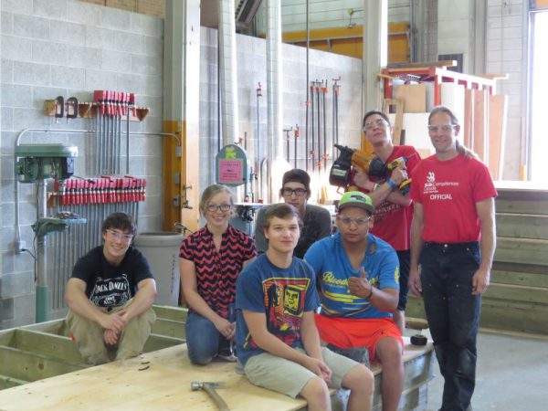 Image shows seven individuals sitting or standing near a wood platform, with carpentry tools around them.