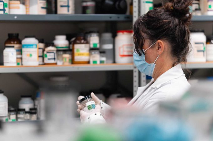 An image of a female wearing a lab coat and mask and looking at medication bottles.
