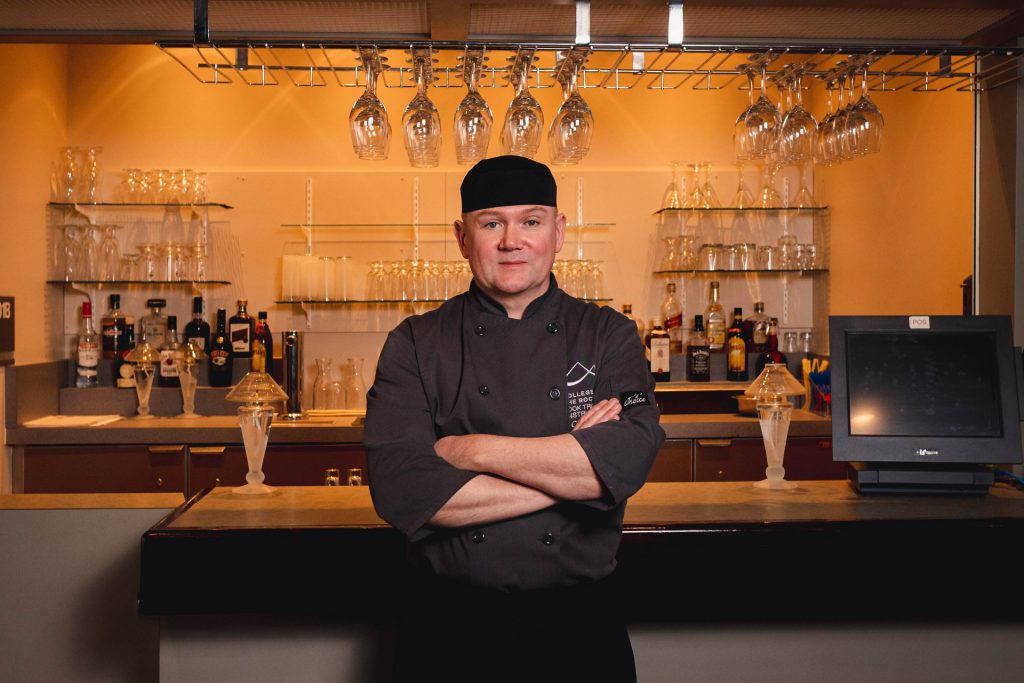 Image shows man in chef's jacket standing with arms crossed in front of a bar.