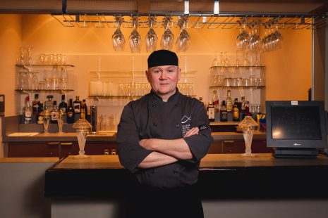 Image shows man in chef's jacket standing with arms crossed in front of a bar.