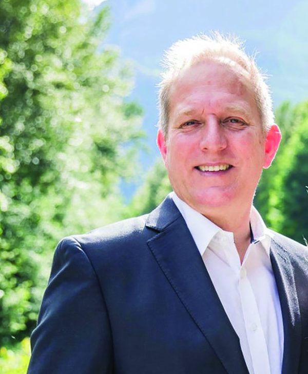 Image shows man in a suit standing in front of a mountain and trees