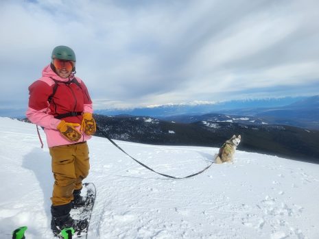 Image shows man on a snowboard atop a mountain with a husky on a leash.