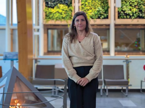 Image shows woman with long brown hair standing beside fireplace.